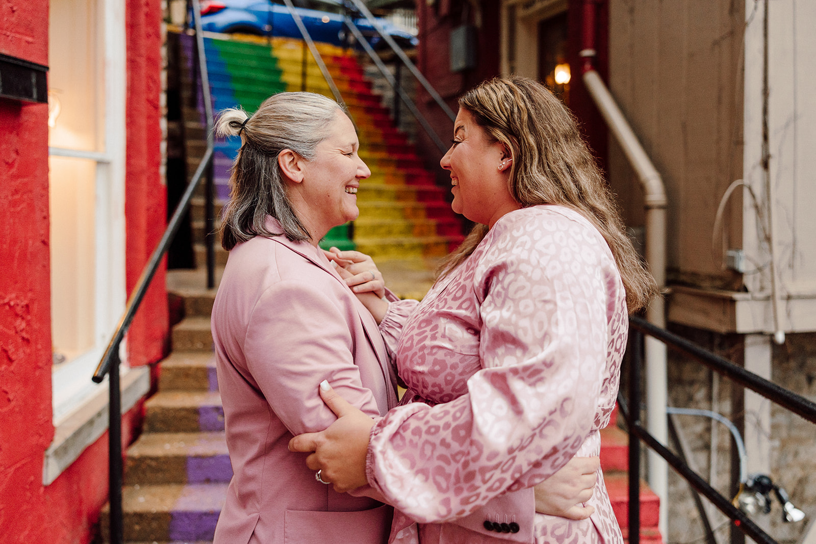 Women smiling at each other in front of rainbow stairs during their city engagment session