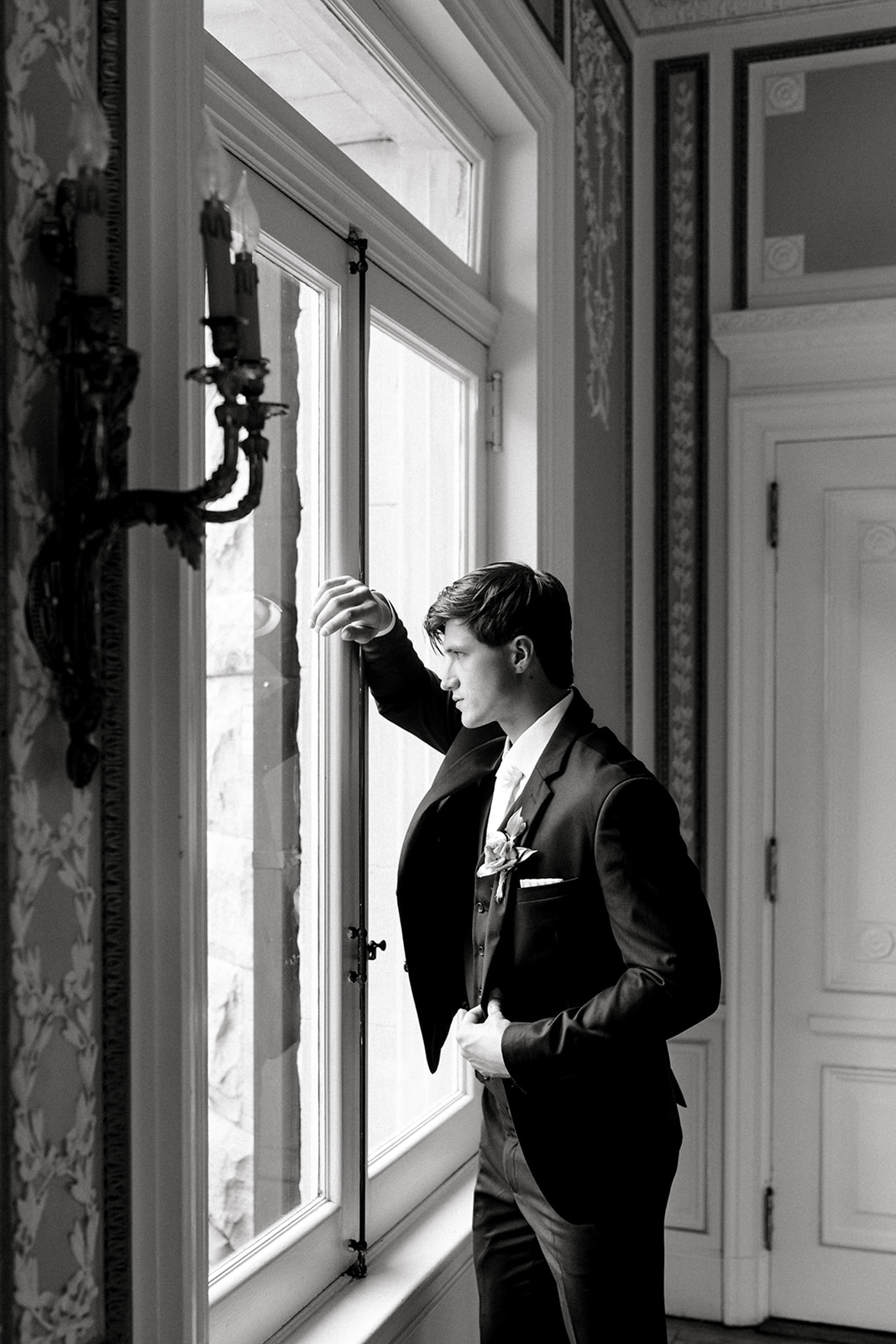 Groom in classic black tuxedo looking out the window, reflecting the luxury wedding attire from The Modern Groom.